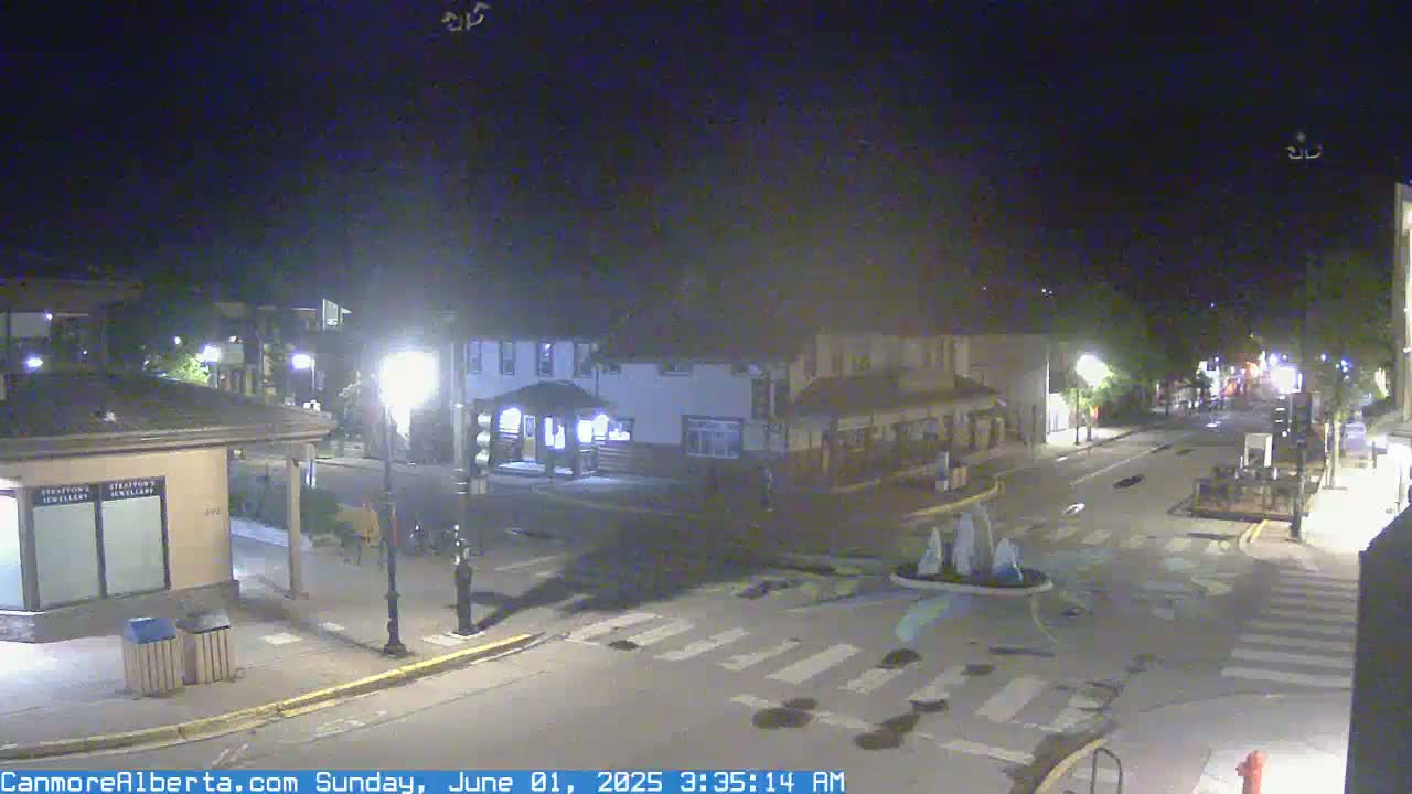A nighttime view of a quiet town square with a crosswalk and a small fountain, under a clear, dark sky.