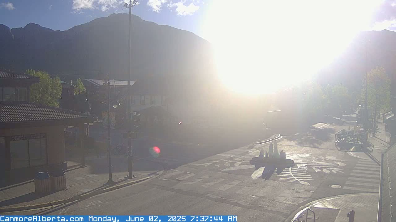 A hazy sunrise illuminates a town square with buildings and trees, mountains in the background.