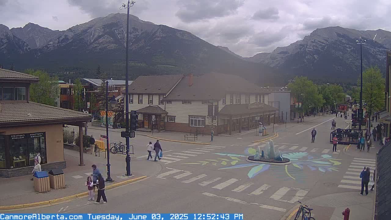 A street scene in a mountain town on an overcast day shows pedestrians crossing a street intersection with painted designs, buildings lining the street, and mountains in the background.