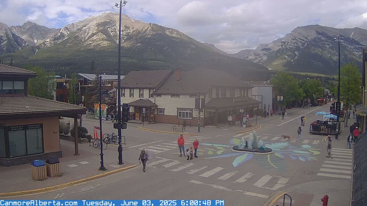 A town square with a painted crosswalk and fountain, surrounded by buildings, under a cloudy sky with mountains in the background.