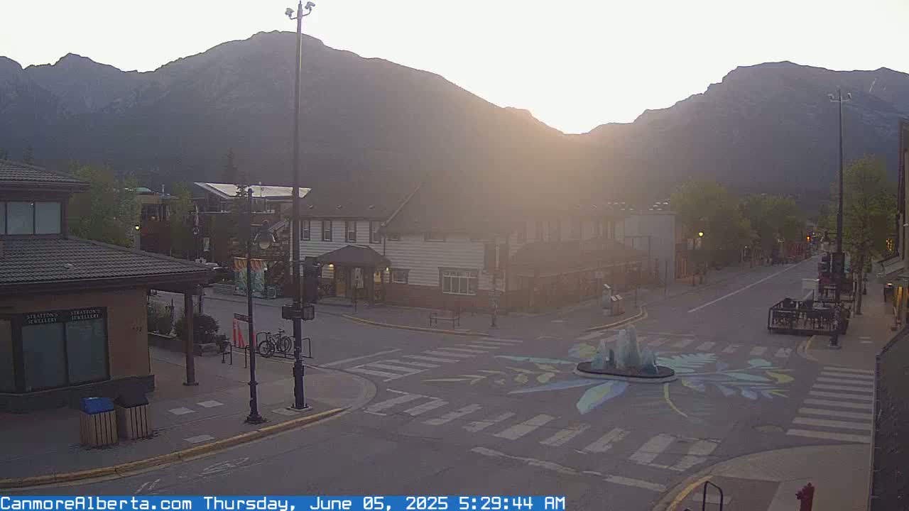 A hazy sunrise illuminates a mountain-backed town square with a painted crosswalk and a small fountain at its center.