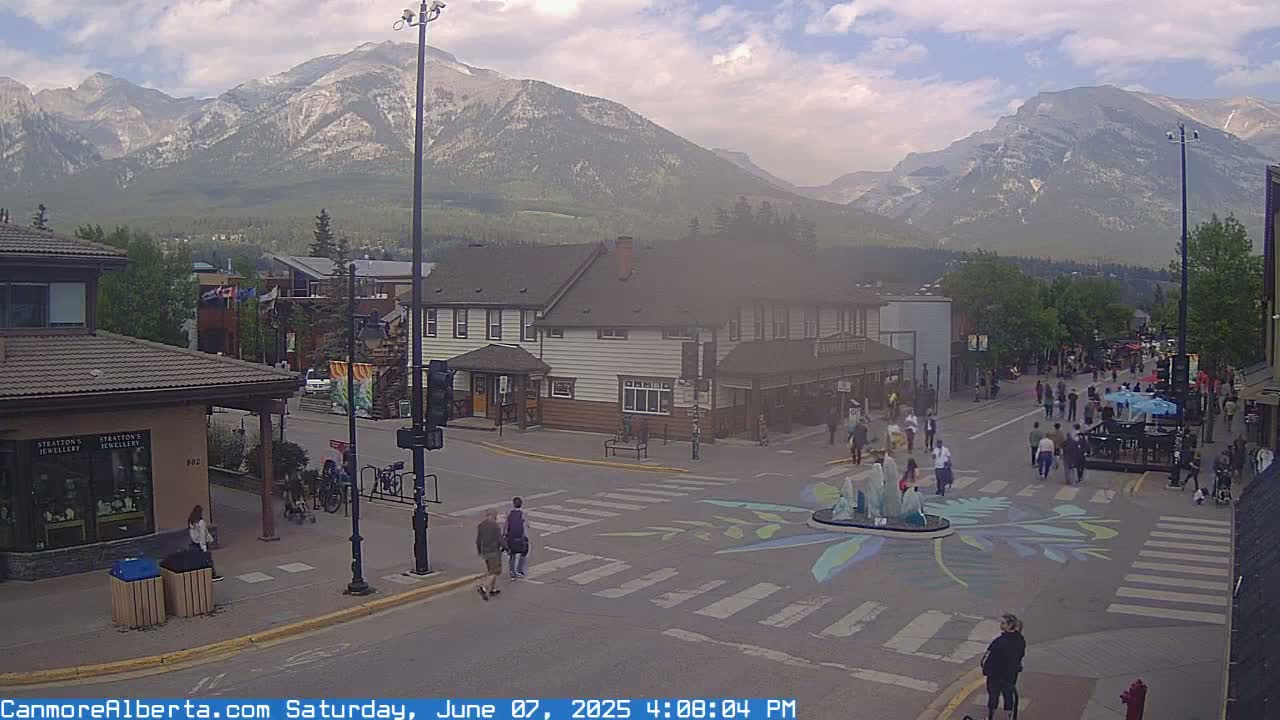 A hazy, partly cloudy day reveals a mountain range in the background of a town square, where many people are walking around a crosswalk with painted designs near a fountain.