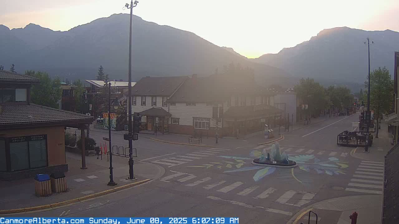 A hazy sunrise illuminates a quiet town square nestled against a backdrop of mountains.