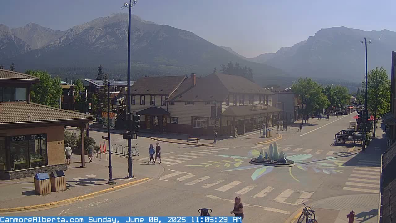 A sunny street scene in a mountain town features a crosswalk with a painted design around a fountain, buildings, pedestrians, and bicycles, all under a hazy sky.