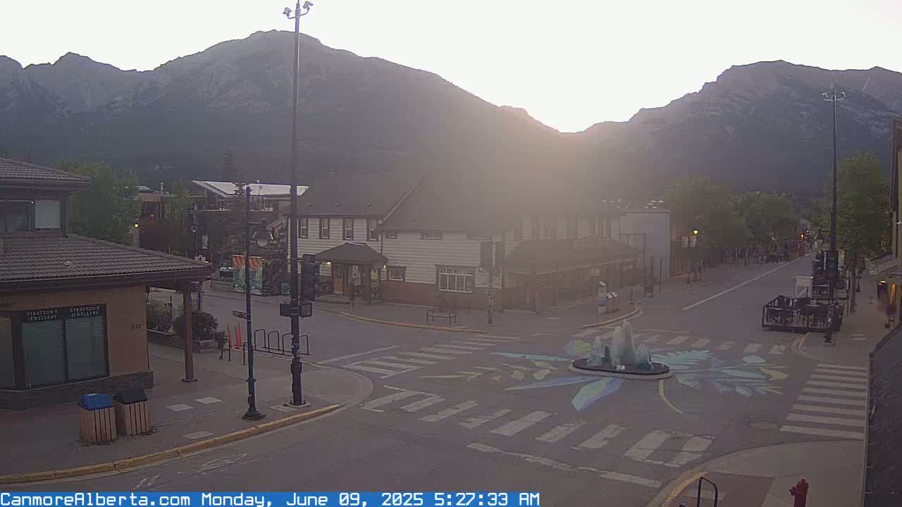 A hazy sunrise illuminates a mountain town street scene with a small fountain at a crosswalk, showing some buildings and a few people.