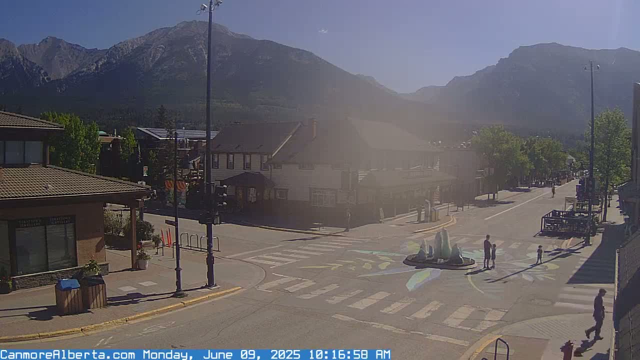A sunny street scene in a town nestled against a mountain range, showing a crosswalk, a small fountain, and a few pedestrians.