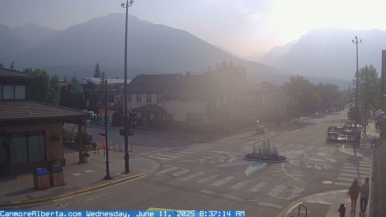 A hazy sunrise illuminates a town square with a crosswalk, buildings, and distant mountains under a smoky sky.
