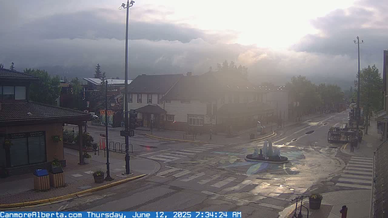 A hazy sunrise illuminates a town square with a small fountain at its center, bordered by low buildings and trees under a mostly cloudy sky.