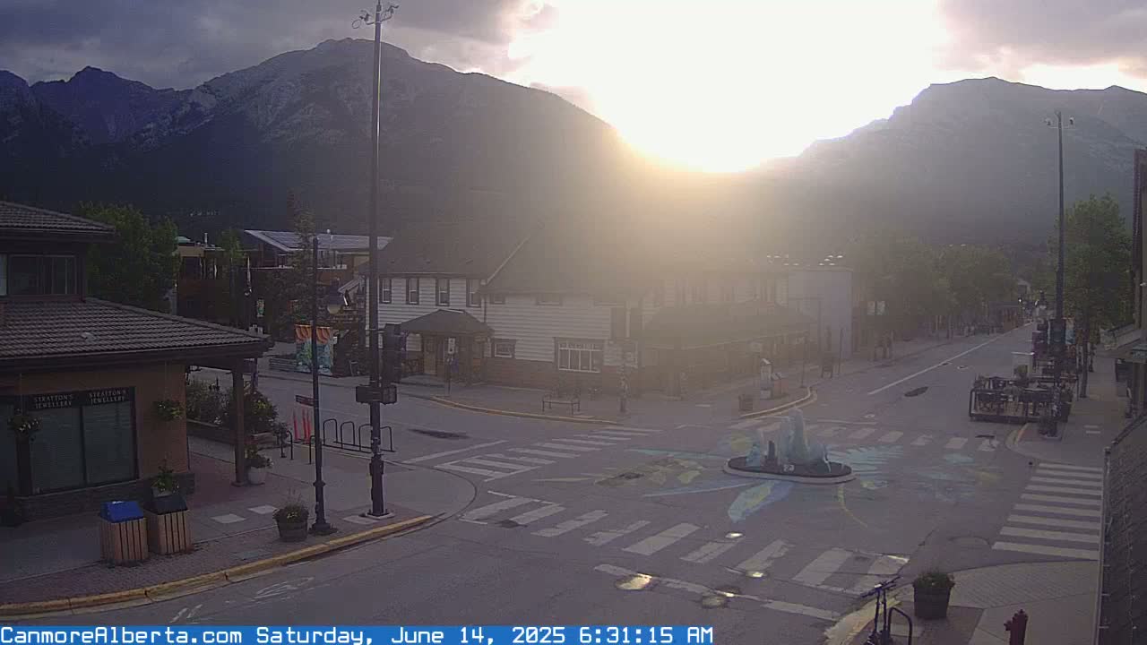 A hazy sunrise illuminates a mountain town street scene with a crosswalk featuring a painted design around a fountain, under partly cloudy skies.