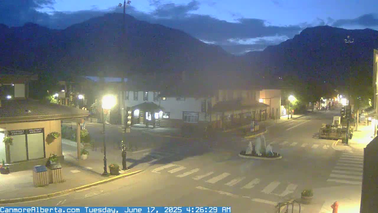 A mostly empty town square at night, under a partly cloudy sky, is lit by streetlights, with mountains visible in the background.