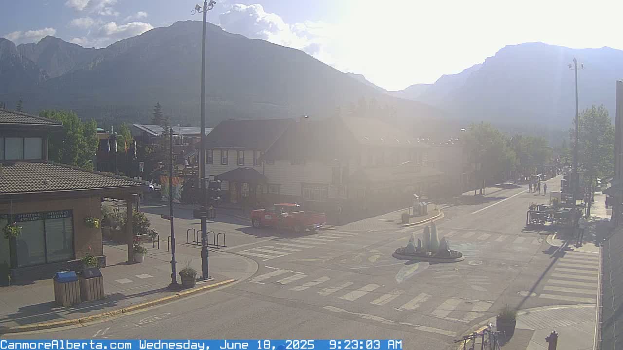 A sunny, hazy day in a mountain town shows a street intersection with buildings, a small fountain, and a red truck, with mountains in the background.
