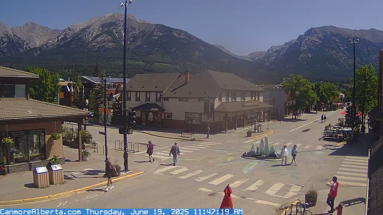 A sunny mountain town square with pedestrians crossing the street, a small fountain, and shops, all under a clear sky.