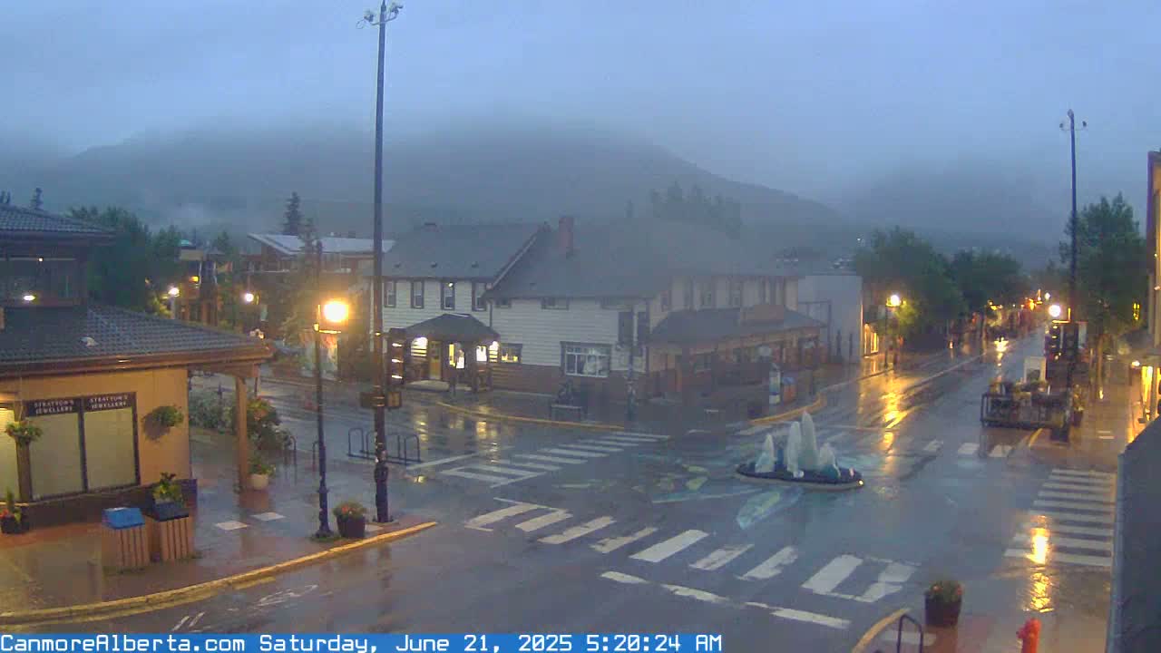 A rain-soaked town square, with low-lying fog obscuring the mountains in the background, shows a crosswalk and a small fountain in the center.