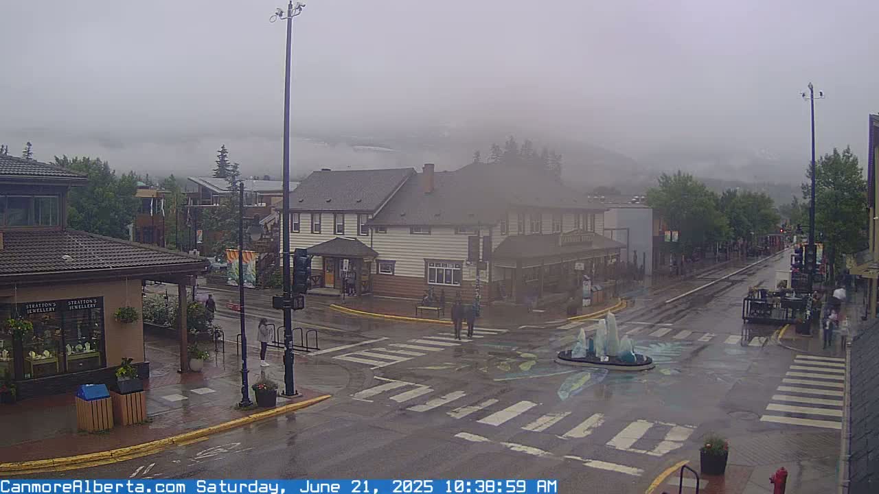 A foggy, rainy street scene shows a town square with pedestrians and buildings, including a multi-story structure, situated in front of a mountain shrouded in mist.