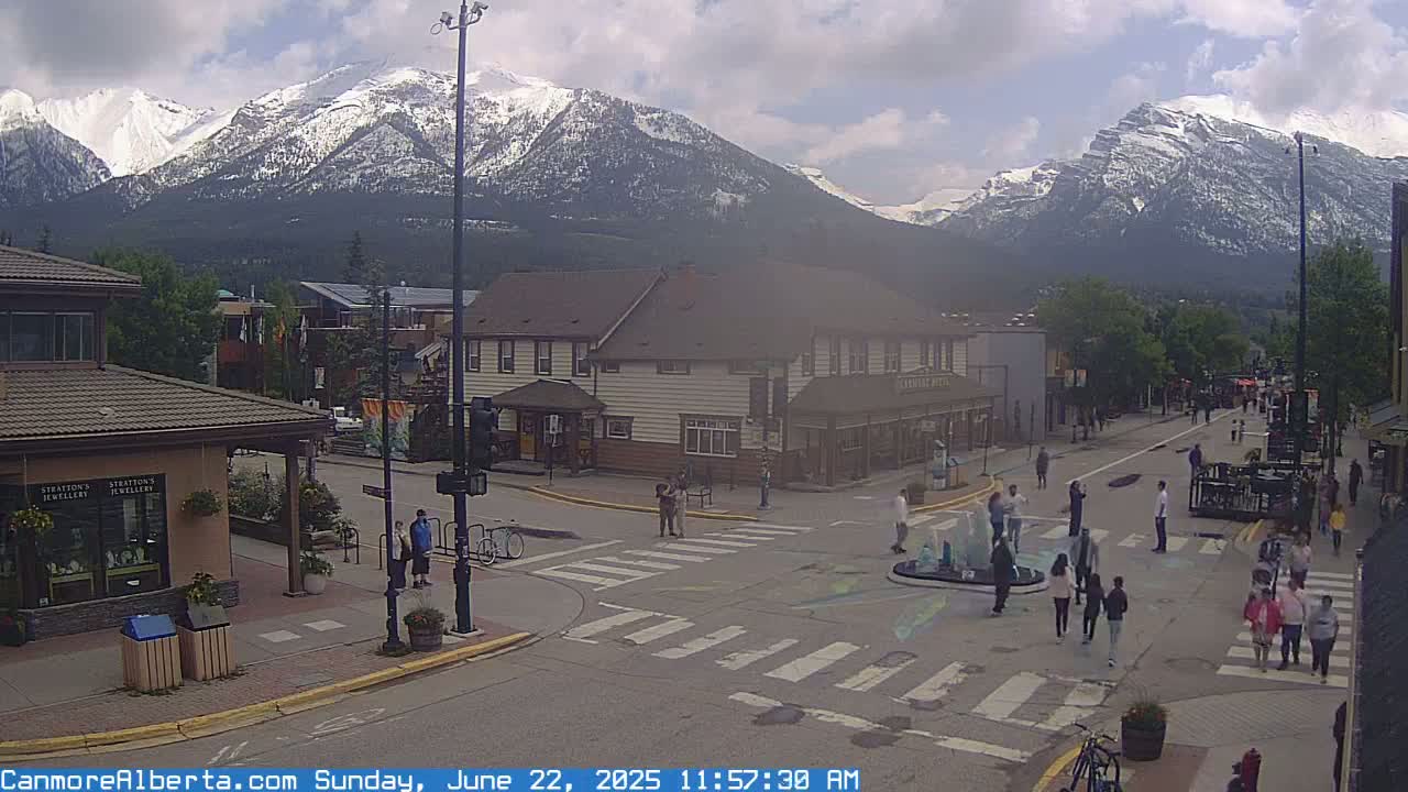 A partly cloudy day shows a town square with people walking around a fountain, bicycles parked on the street, and snow-capped mountains in the background.