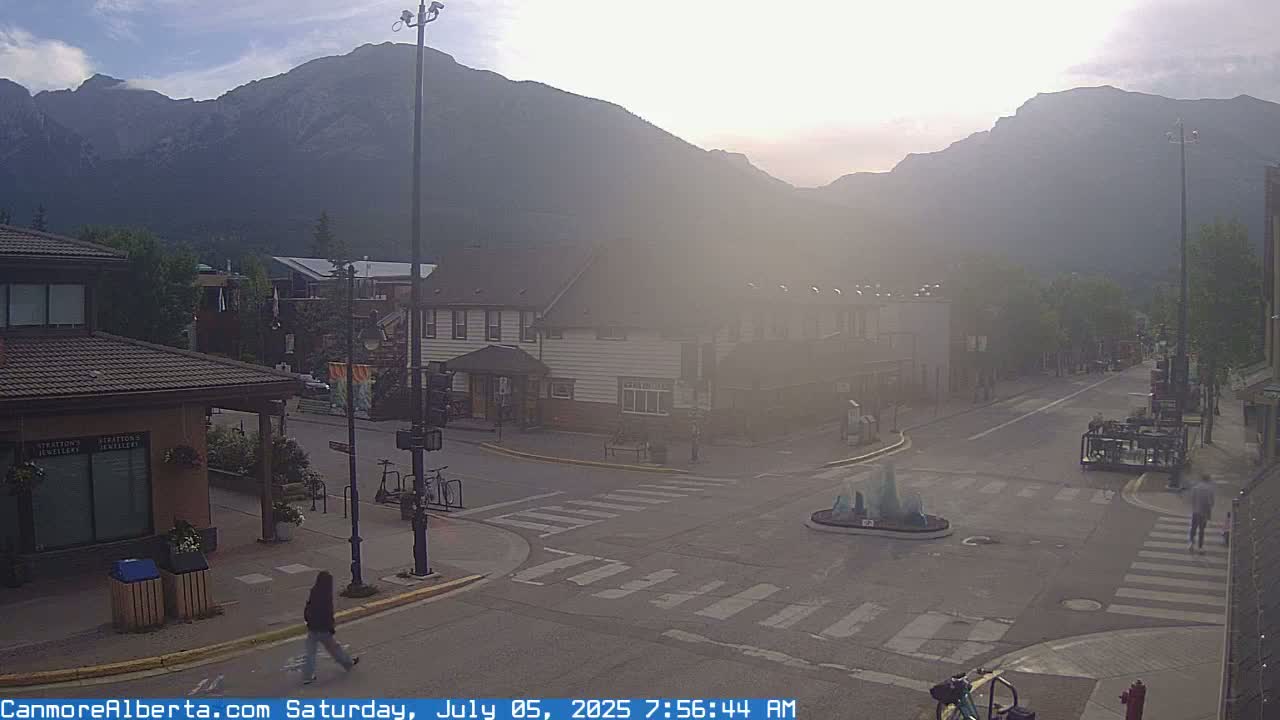 A hazy sunrise illuminates a mountain-backed town square with a fountain at its center, where a few pedestrians walk on the street.