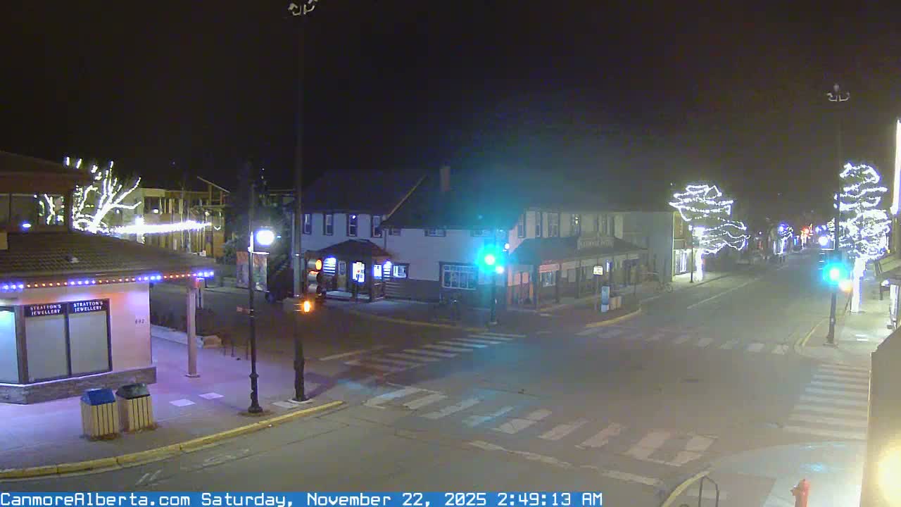 On a clear, dark night, a quiet urban street intersection is illuminated by festive white string lights on trees and buildings, with dry roads and active traffic signals visible.