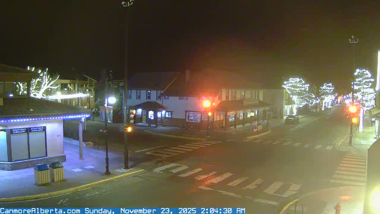 A quiet town intersection is seen at night, illuminated by streetlights and numerous trees wrapped in bright white holiday lights, under hazy conditions.