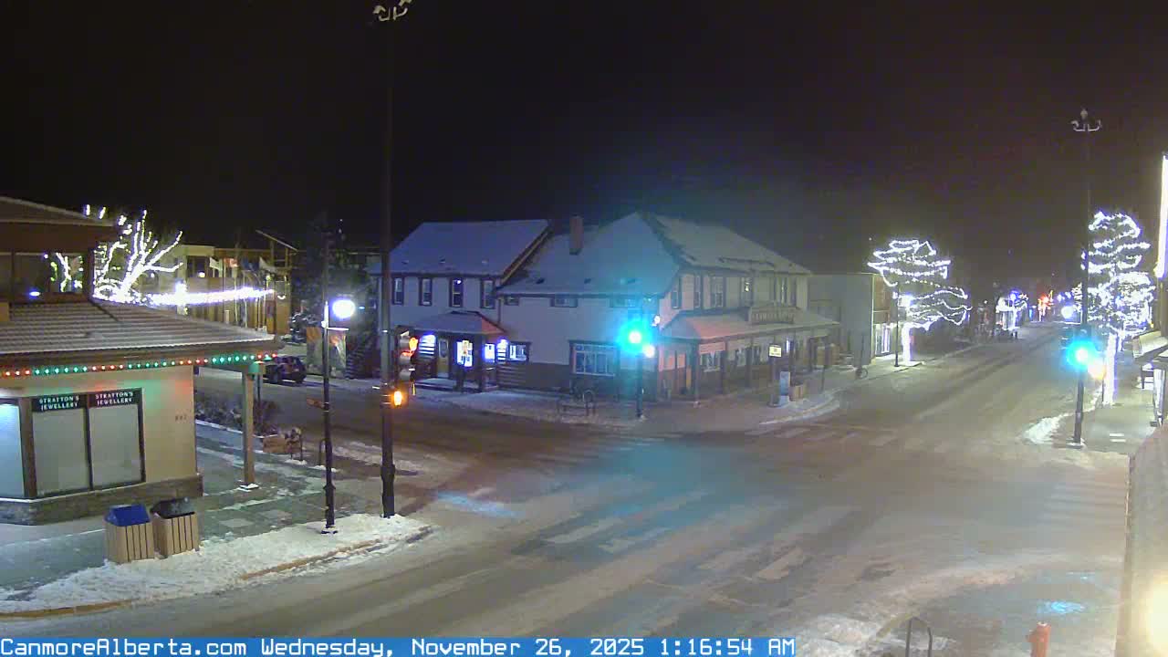 A quiet, snow-covered street in a town glows at night, illuminated by streetlights, festive white string lights on trees and buildings, and an intersection showing green and amber traffic signals.