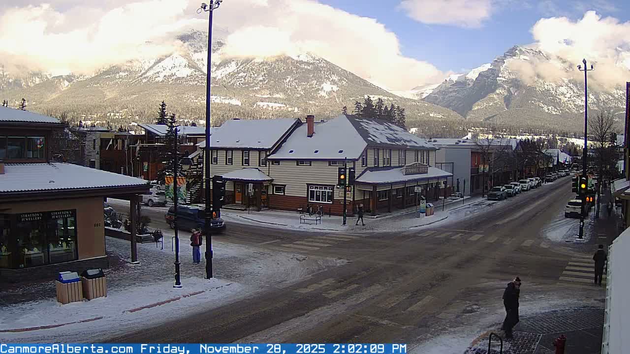A snowy town street scene features several buildings with snow-covered roofs and a street with slushy patches, with a few pedestrians and parked cars, all set against majestic, snow-capped mountains partially obscured by clouds under a bright, clear-ish sky.