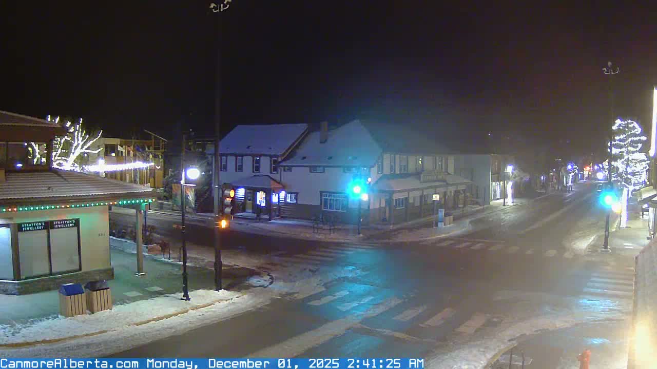 A clear, cold winter night scene shows a quiet, snow-covered urban intersection with buildings and trees decorated with festive lights, all illuminated by streetlights and active traffic signals.