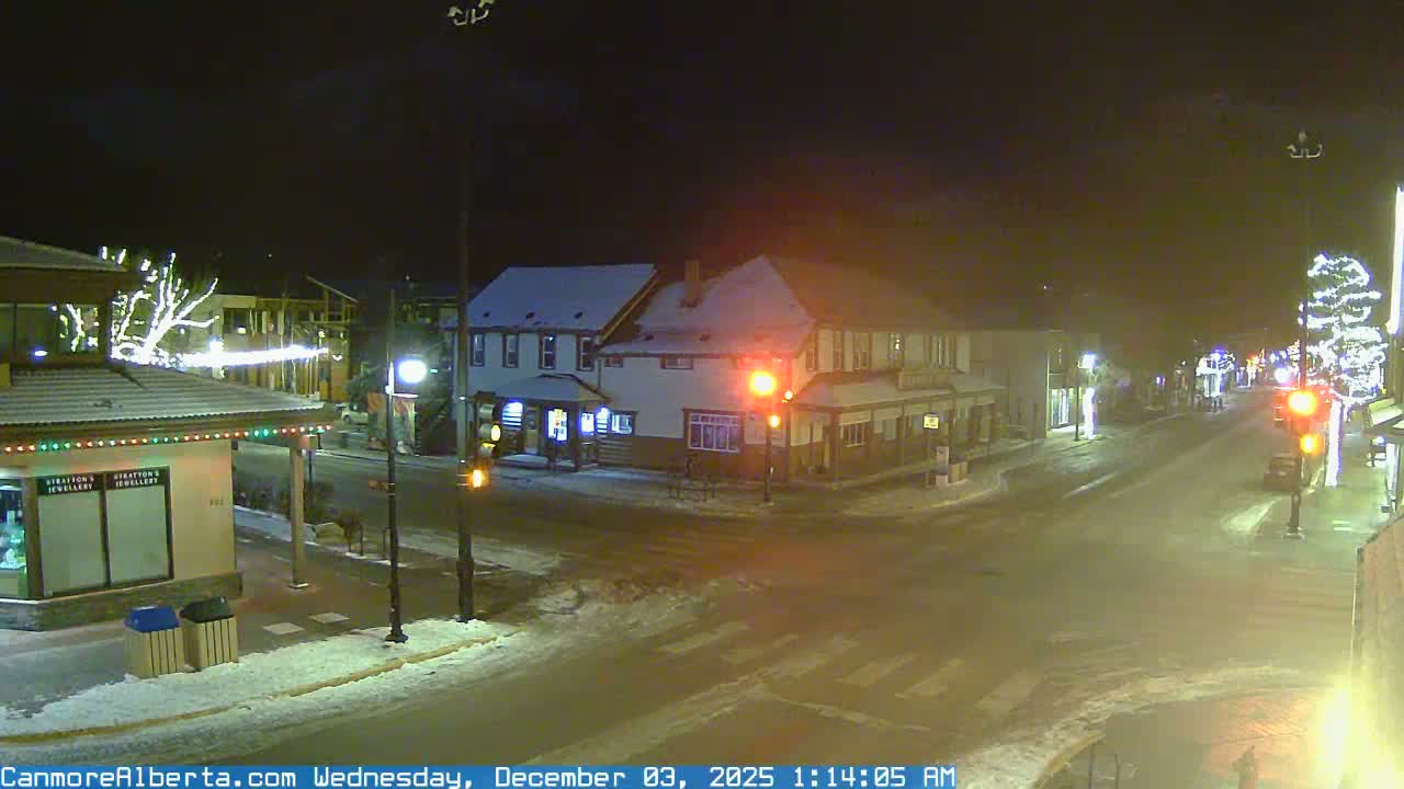 A snow-covered street in a town is illuminated by festive holiday lights, streetlights, and traffic signals at night, indicating cold, clear winter weather.