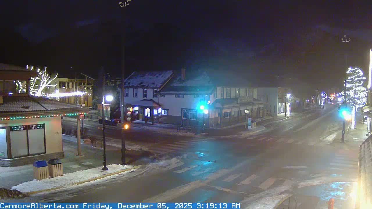 A snow-dusted town street at night is illuminated by streetlights, traffic lights, and festive holiday lights, under clear, cold winter conditions.