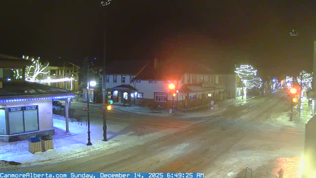 A snow-dusted town street at night is illuminated by streetlights, traffic lights, and festive holiday lights, under clear, cold winter conditions.