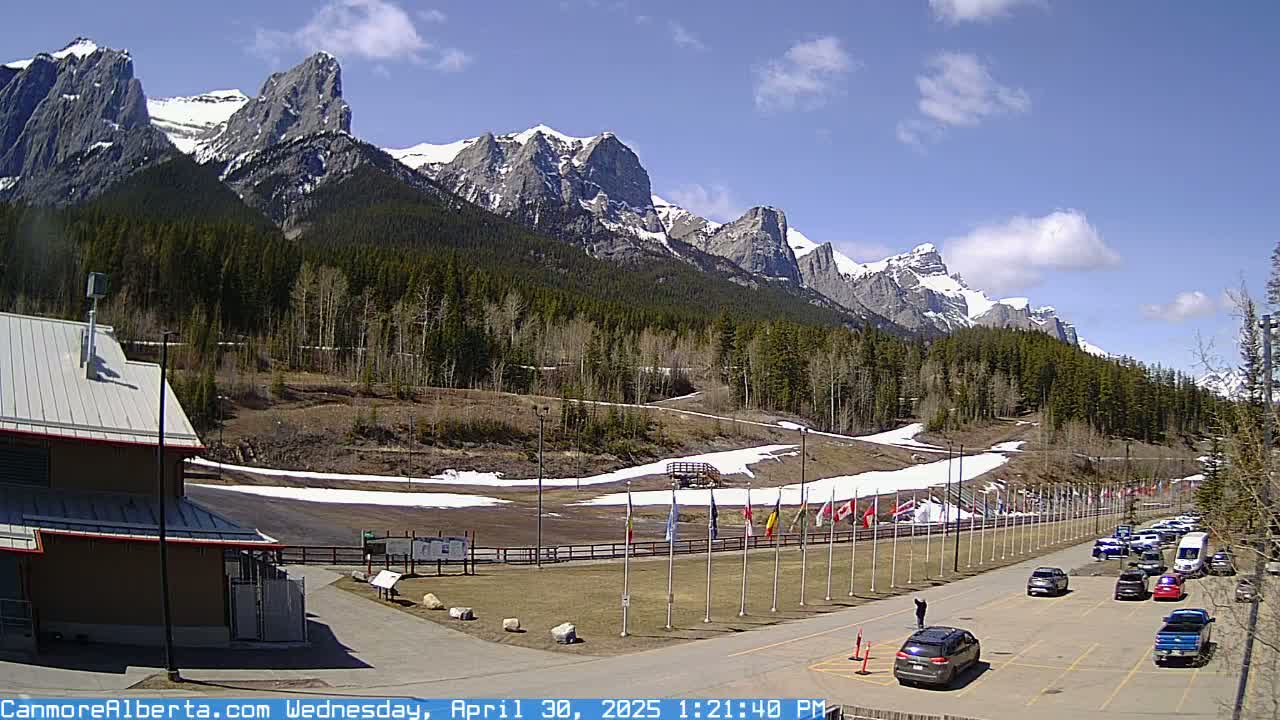 A parking lot with several cars sits in front of a grassy area with snow patches, a wooden structure, and a row of flags, all nestled at the base of a snow-capped mountain range under a mostly sunny sky.