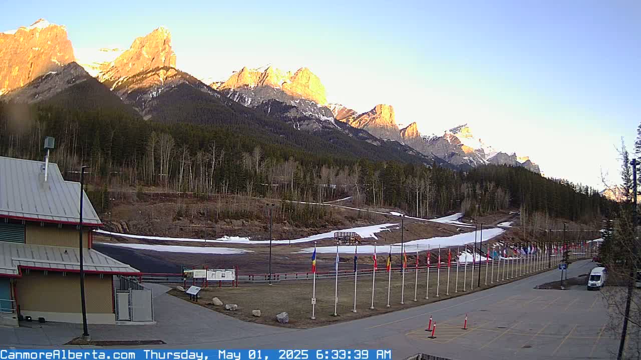 A sunrise illuminates a mountain range behind a paved area with patches of snow and several flags lined up along a road, next to a building.