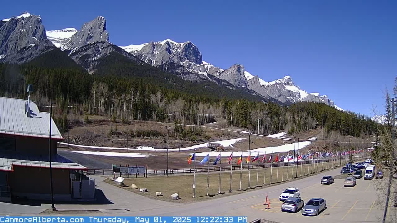 A parking lot with cars is situated in front of a grassy area with snow patches and a line of flags, all set against a backdrop of snow-capped mountains under a clear blue sky.
