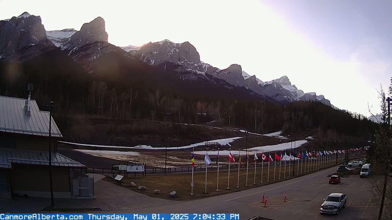A row of flags lines a paved road in front of a building, with a mountainous landscape in the background under a clear, bright sky.