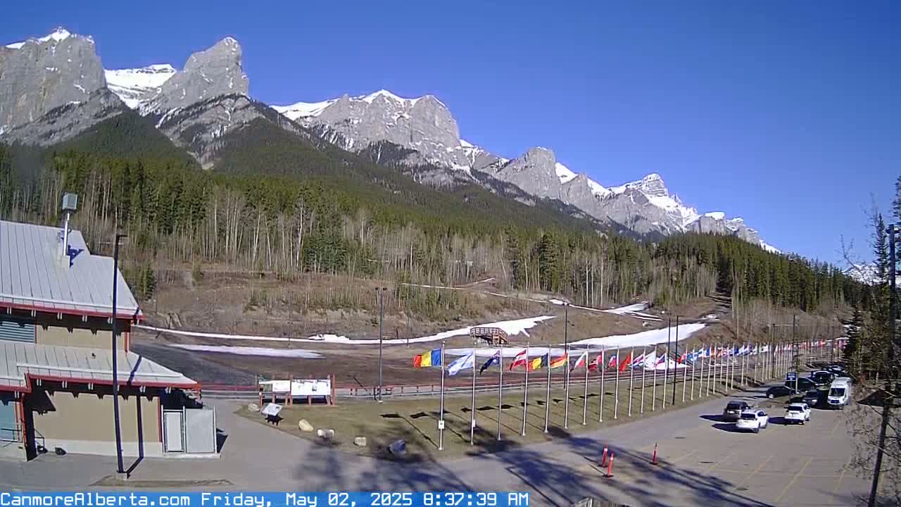 A sunny day reveals a parking lot, a building, and a snow-dusted track lined with numerous flags, all set against a backdrop of snow-capped mountains and a forested hillside.