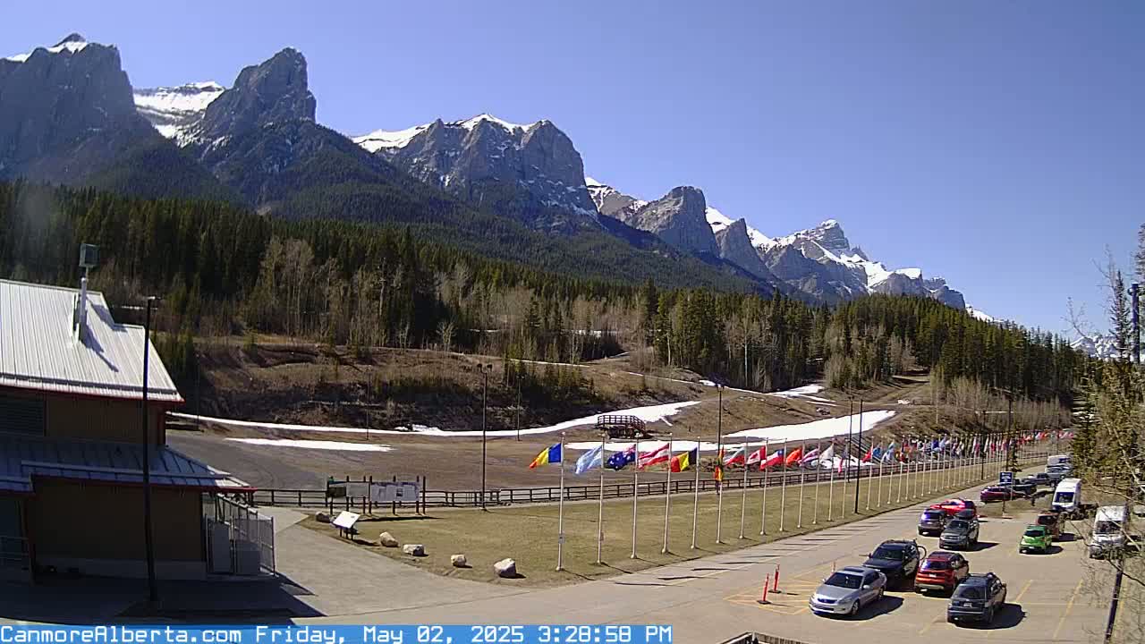A sunny day reveals a mountain range capped with snow behind a paved area with parked cars, a building, and a line of flags on poles.