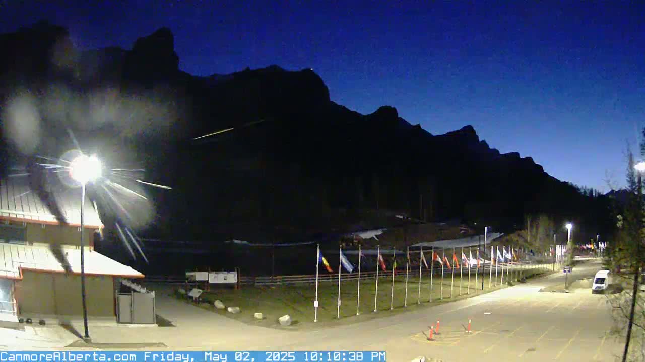 A nighttime view shows a paved road leading toward a row of flags in front of dark mountains under a clear, dark blue sky.