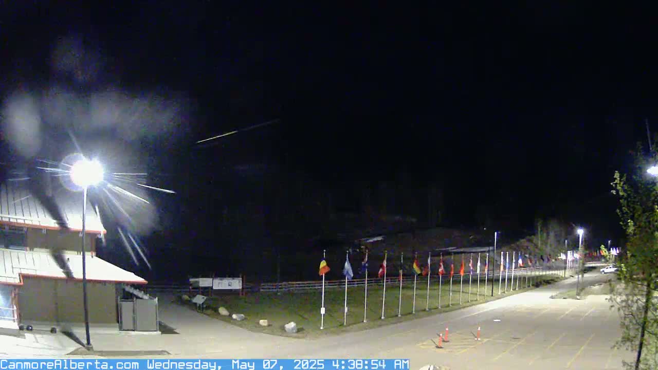 A nighttime scene shows a building, a row of flags, and a mostly empty parking lot under a dark sky with streaks of light possibly from a meteor shower.