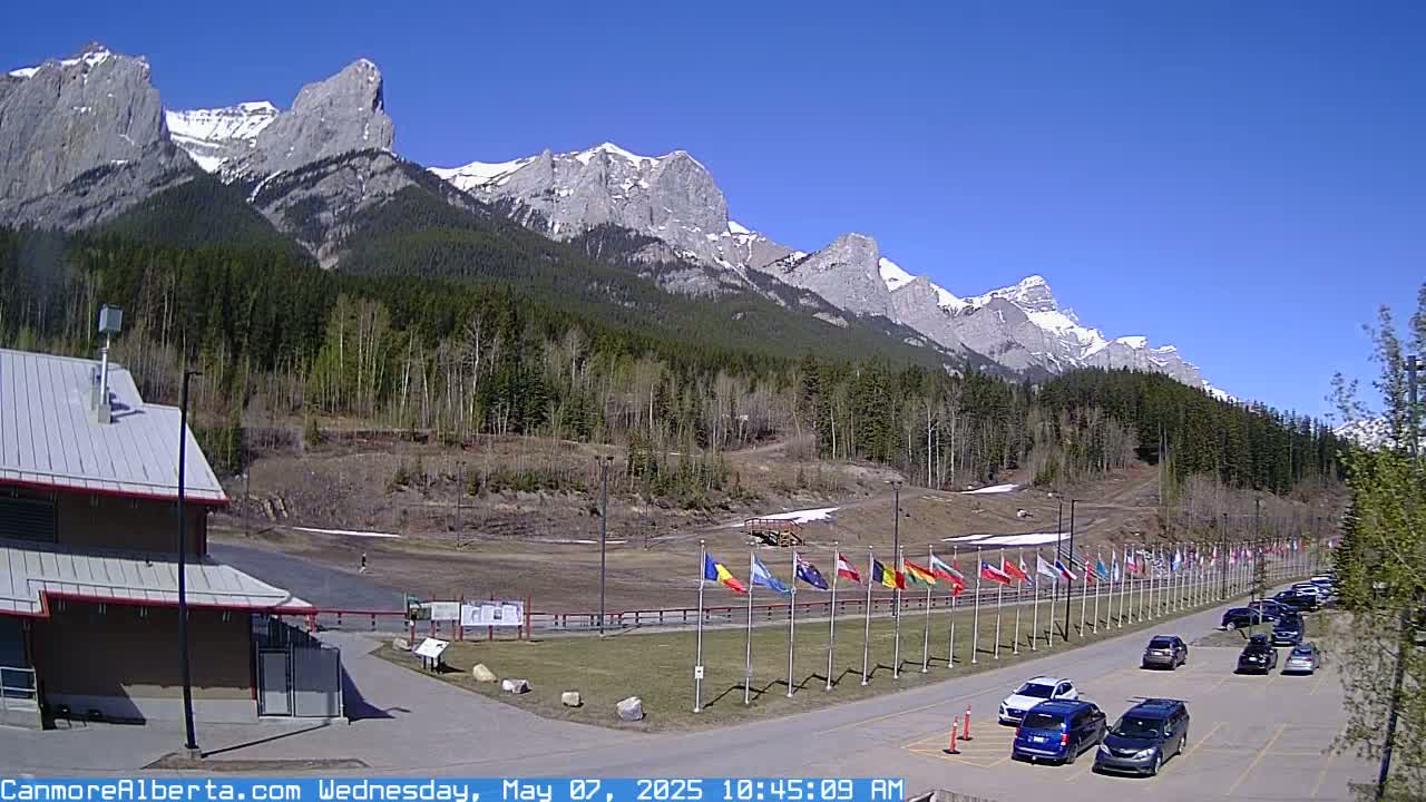 A sunny day reveals a mountain range with patches of snow, partially visible behind a flat, open area with a parking lot, several national flags, and a building.