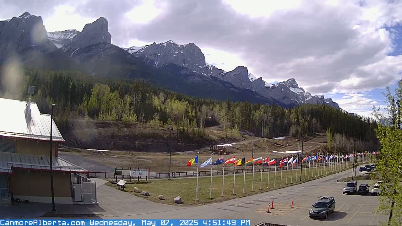 A partially cloudy day reveals a parking lot with several cars, a row of flags, and a building near a forested area with snow-capped mountains in the background.