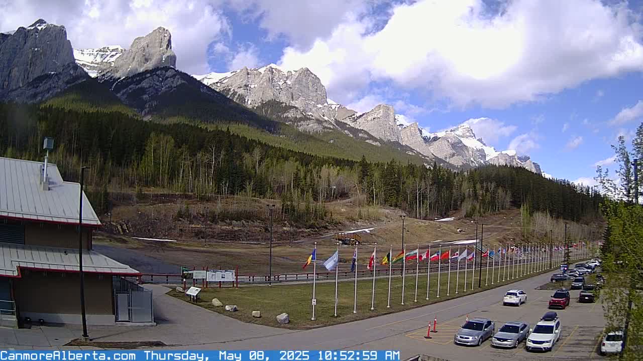 A parking lot with numerous cars is situated in front of a line of flags, overlooking a grassy field and a background of snow-capped mountains under a partly cloudy sky.