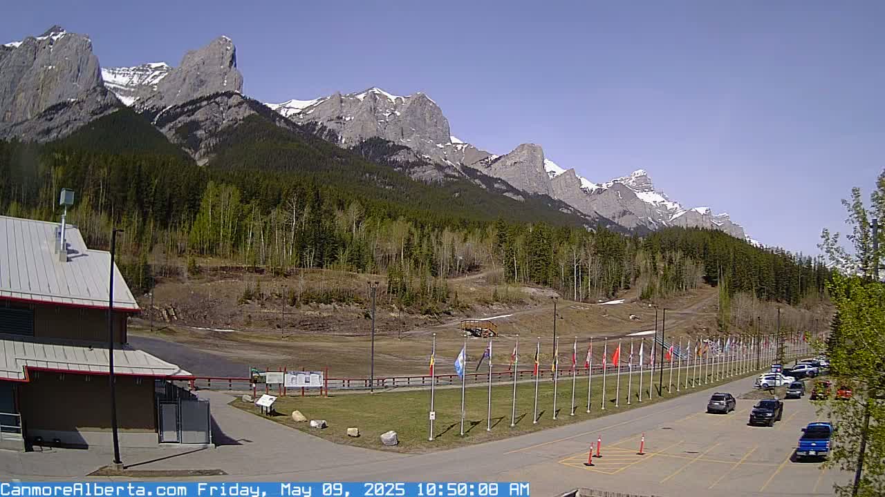 A parking lot with several cars is situated in front of a building, with a row of flags, and a forested mountain range with some snow visible in the background under a clear, sunny sky.