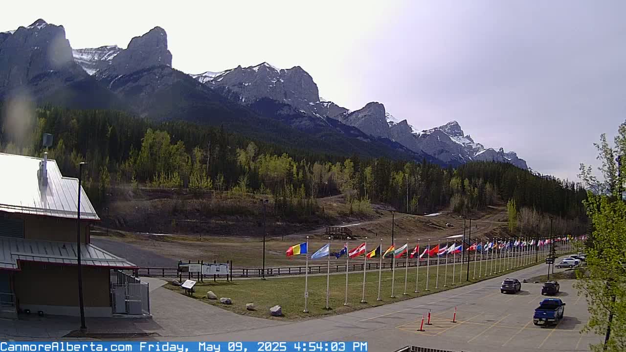 A row of flags stands in a parking lot next to a building, overlooking a forested valley and a mountain range partially covered in snow under a partly cloudy sky.
