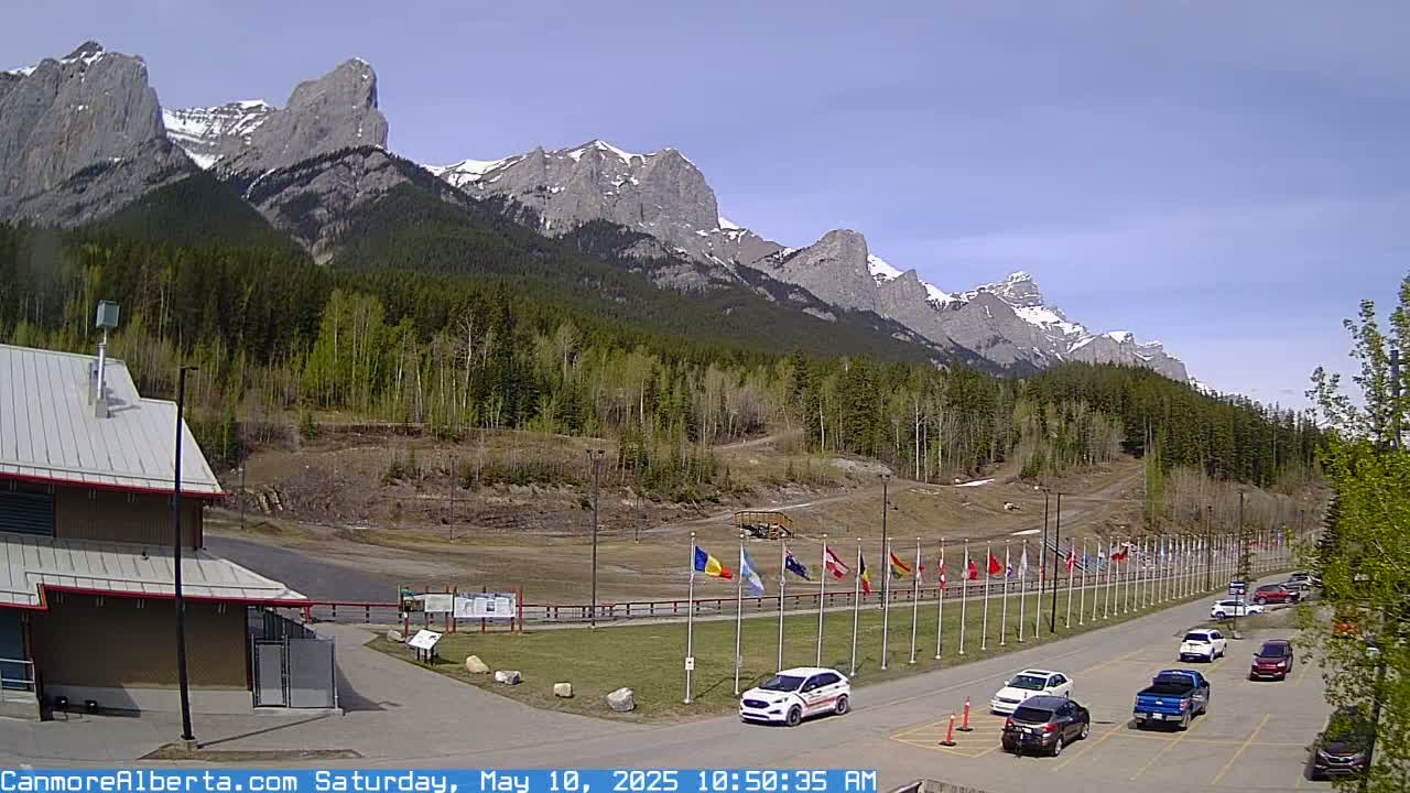 A parking lot with several cars is situated in front of a building, with a line of flags and a mountain range in the background under a clear, sunny sky.