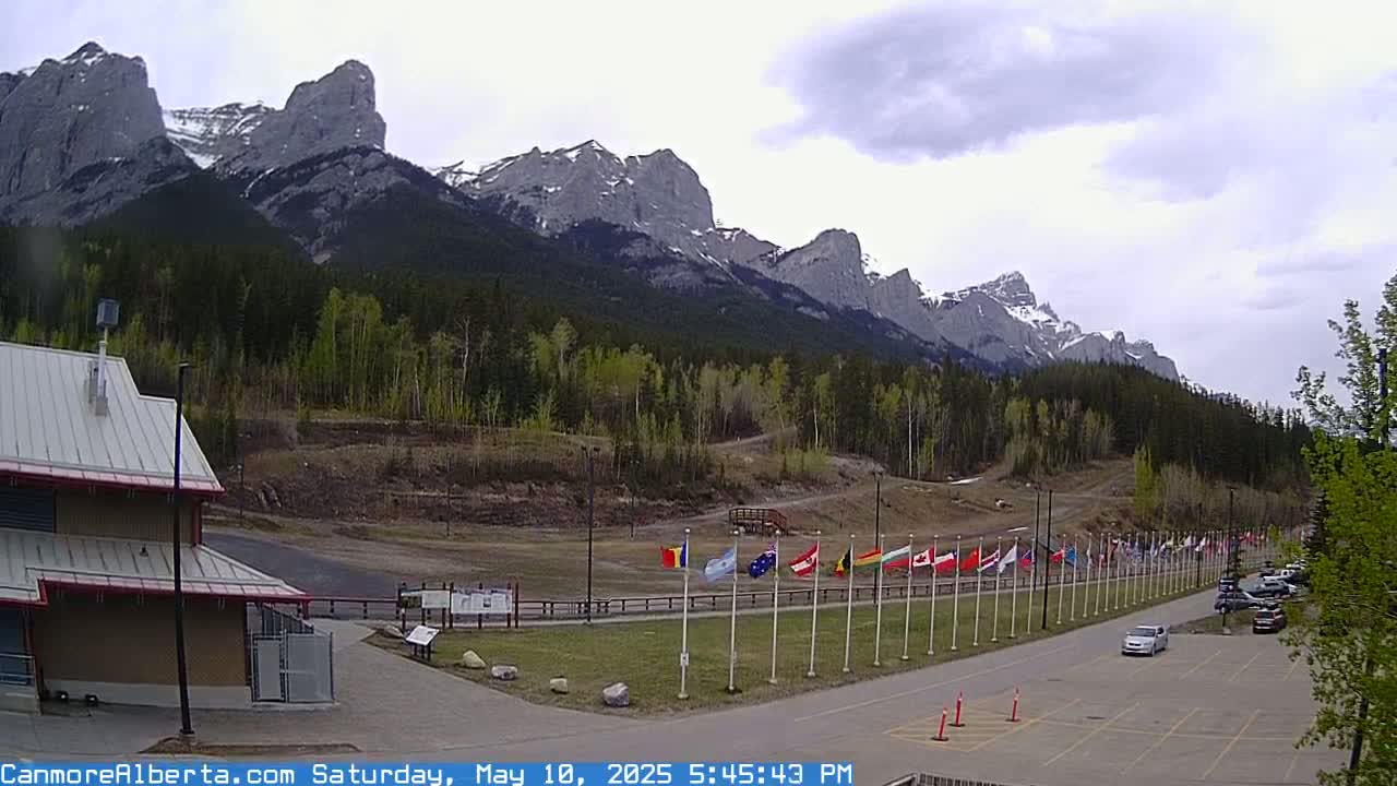 A row of flags stands in front of a building and parking lot, with a mountain range in the background under a cloudy sky.