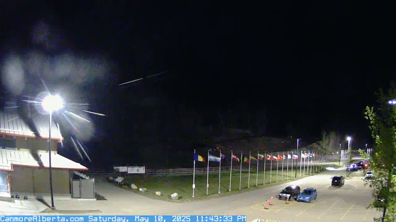 A nighttime scene shows a parking lot with several cars, a building with lights, and a row of flags under a dark sky with streaks of light possibly from meteors.
