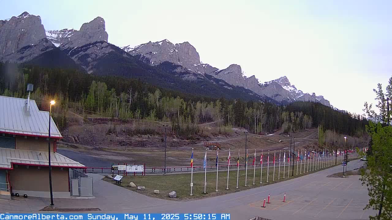 A row of flags lines a paved path leading to a building near a mountain range under a clear, light-grey dawn sky.