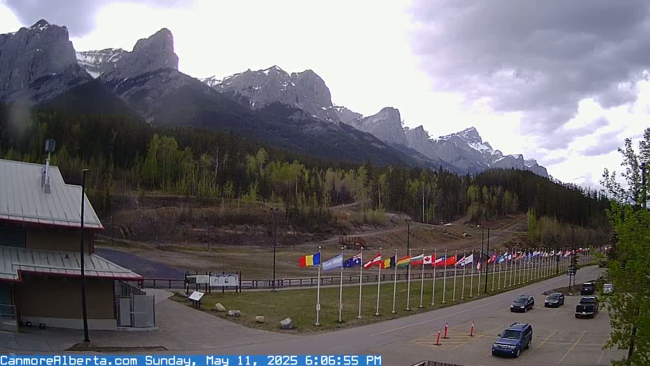 A parking lot with several cars sits before a row of flags, which are in front of a forested area with a mountain range in the background under a partly cloudy sky.