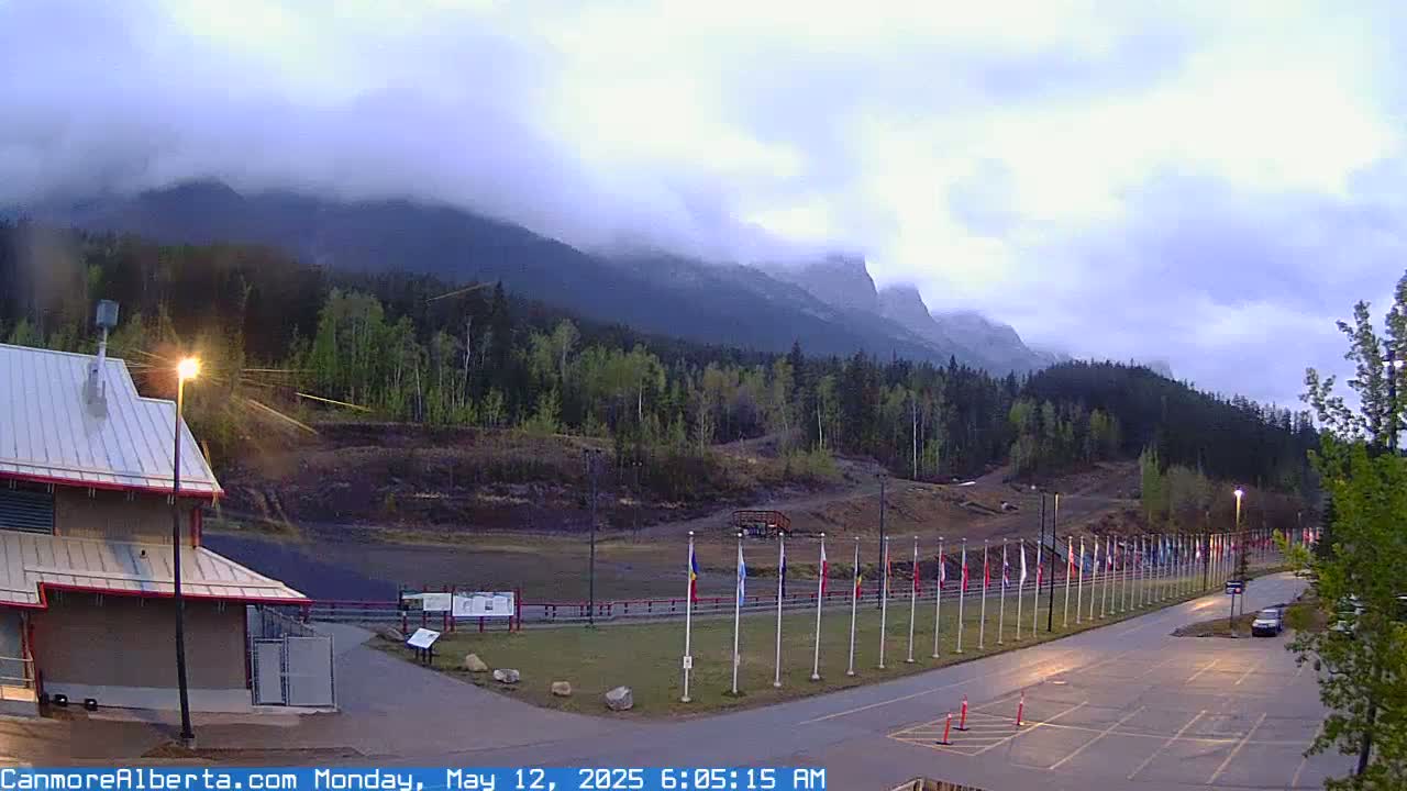 A mostly cloudy morning scene shows a row of flags in front of a building and a forested mountain range in the background.