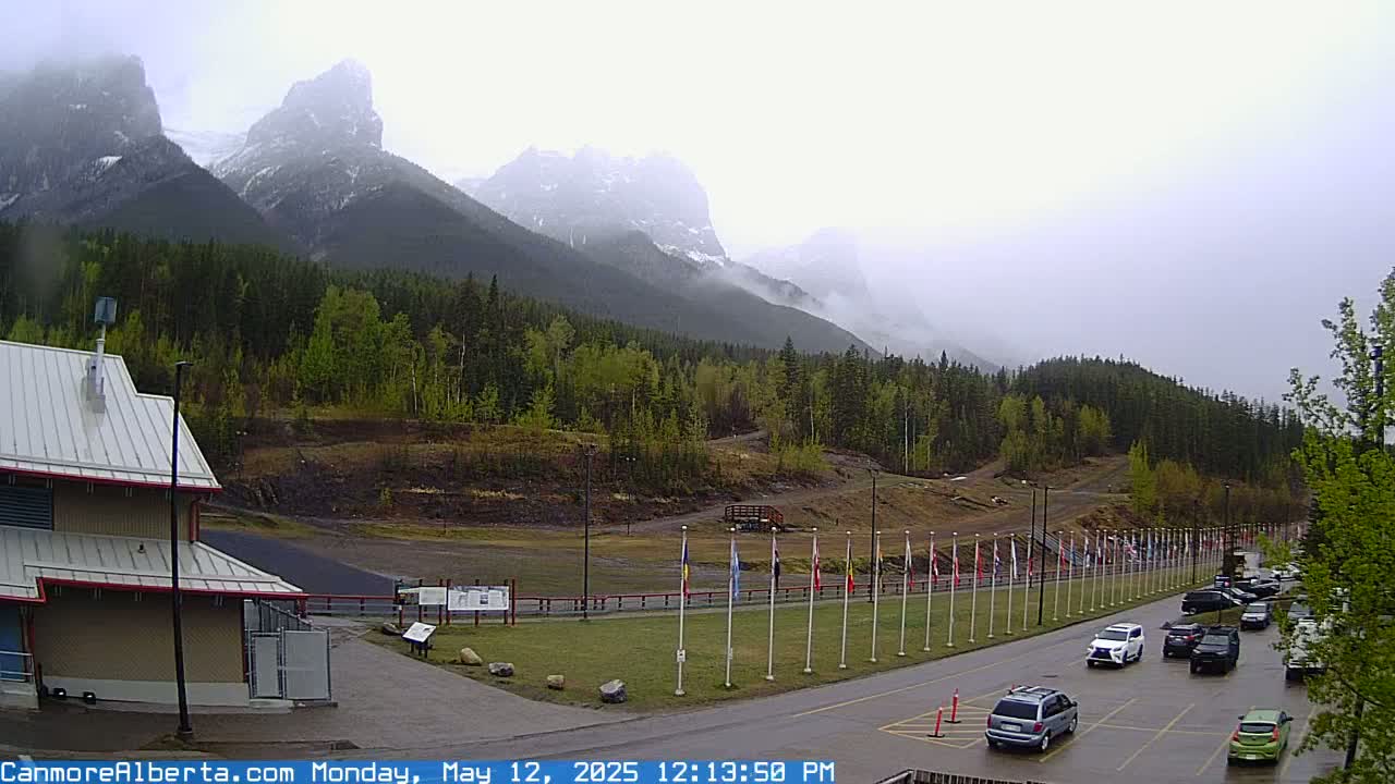 A cloudy day reveals a mountain range partially obscured by mist, overlooking a paved road with parked cars and a line of flags in front of a building and grassy area.