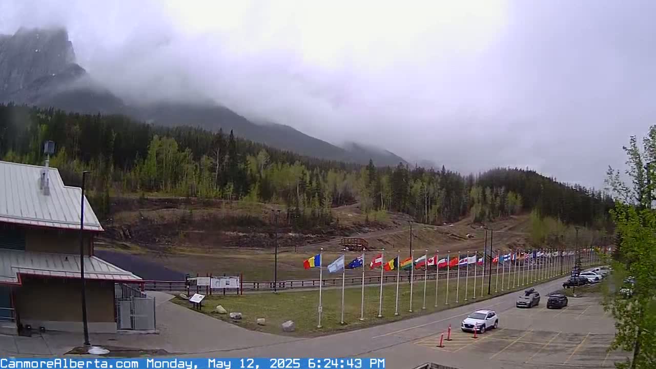 A parking lot with several cars, a row of flags, and a building are situated in front of a mountain range partially obscured by fog.