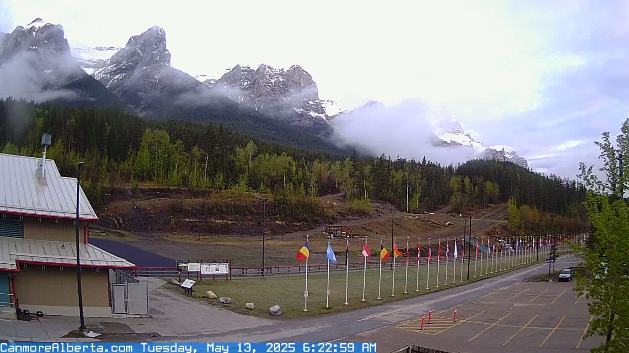 A row of flags lines a paved road leading to a building and parking lot, with misty mountains and a forested hillside in the background.